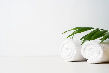 Rolgordijnen Spa-concept: twee witte pluizige handdoeken gedraaid in rollen op een lichte ondergrond met een palmblad met kopieerruimte © PINKASEVICH