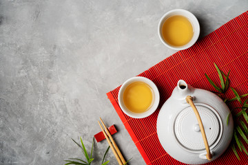 Asian food background with tea set and chopsticks on red bamboo mat on gray stone background. Top...