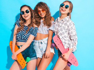 Three young stylish smiling beautiful girls with colorful penny skateboards. Women in summer hipster checkered shirt clothes posing near blue wall in studio in sunglasses. Positive models having fun 