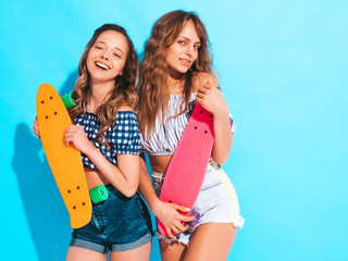 Two young stylish smiling beautiful girls with colorful penny skateboards. Women in summer hipster checkered shirt clothes posing near blue wall in studio. Positive models having fun and going crazy