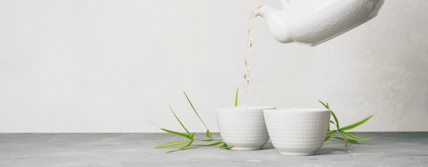 Female hand pouring green tea from a teapot into cups on white background with copy space. Long...