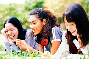 Happy and smiling mixed diverse racial female student friends lying down on grass while using a computer laptop together in park.