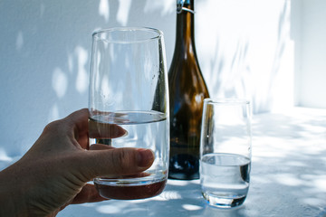 Hand holding a water glass in front of a bottle and another glass, isolated on a white concrete wall with shadows of leaves on it