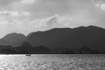 Silhouettes of fishing boats against the backdrop of mountains and a sunny sky with clouds. Black and white photography