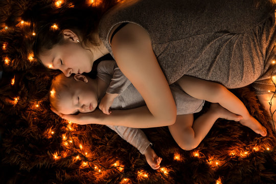 A young mother and her baby are sleeping on a fluffy brown blanket, lights are spread around them