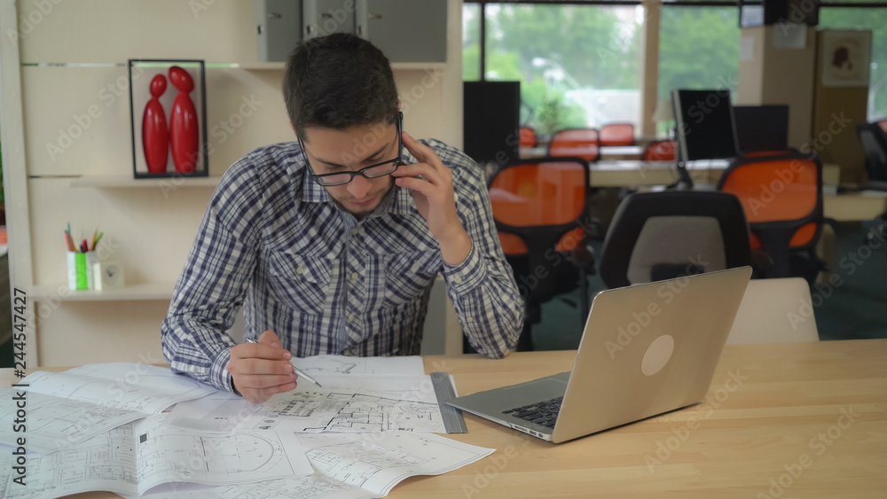 Aufkleber Architect checking details and make notes on the paper plan. Blueprint documents and computer on the wooden table with equipment for drawing and draftsmanship. Male wearing in casual clothes and