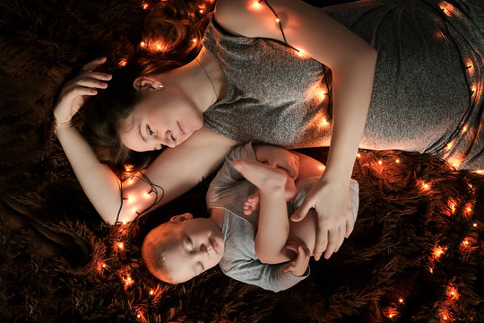 A young mother and her baby are sleeping on a fluffy brown blanket, lights are spread around them