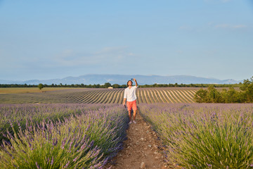 The beautiful brutal young man with long brunette hair makes selfie by cellphone in the field of lavender, white shirt and red shorts