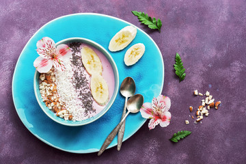 Obraz na płótnie Canvas Fresh healthy breakfast - yogurt with banana, almond,coconut flakes, chia seeds in a blue bowl decorated with flowers on a purple background. Top view