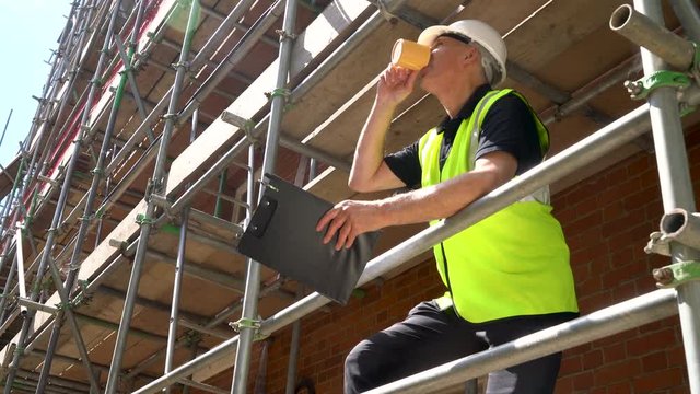 Male builder foreman, worker or architect working on construction building site standing on scaffolding with a clipboard and drinking mug of tea or coffee