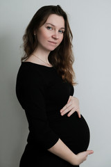 Young pregnant girl in black dress stands on white background and hugs her belly