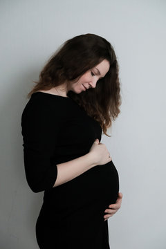 Young pregnant girl in black dress stands on white background and looks at her belly