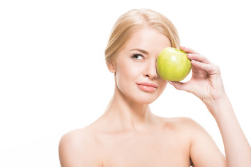 naked blonde girl holding green apple near eye and looking away isolated on white
