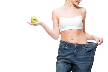 cropped shot of smiling slim woman in oversized jeans holding green apple isolated on white