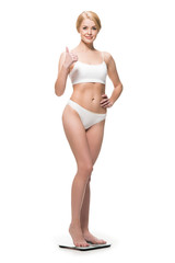 Obraz premium attractive happy slim girl in underwear standing on scales and showing thumb up isolated on white