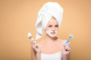 young woman with towel on head and shaving cream on face holding razor and shaving brush isolated...