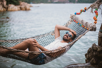 Young man lying in a hammock by the ocean. A handsome guy is resting in a hammock against the backdrop of the mountains by the sea.