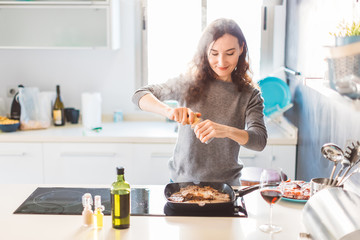 Young smiling woman cooking in the kitchen, adding pepper to the grilled meat. Healthy food concept.