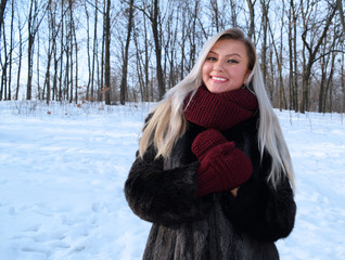 Portrait of a cheerful blonde girl in a black fur coat and gloves on the background of a winter...