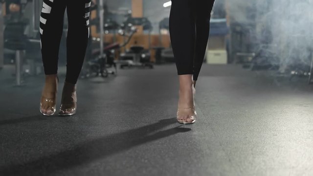 Two fitness bikini blonde girls posing in dark gym in slow motion. Close up view of legs on high heels