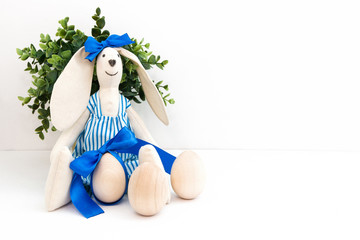 Easter. Handmade easter rabbit bunny sewn from light linen fabric in a striped jumpsuit with a blue bow on the ear. Wooden eggs next to it. White background. Free space for text. Concept.