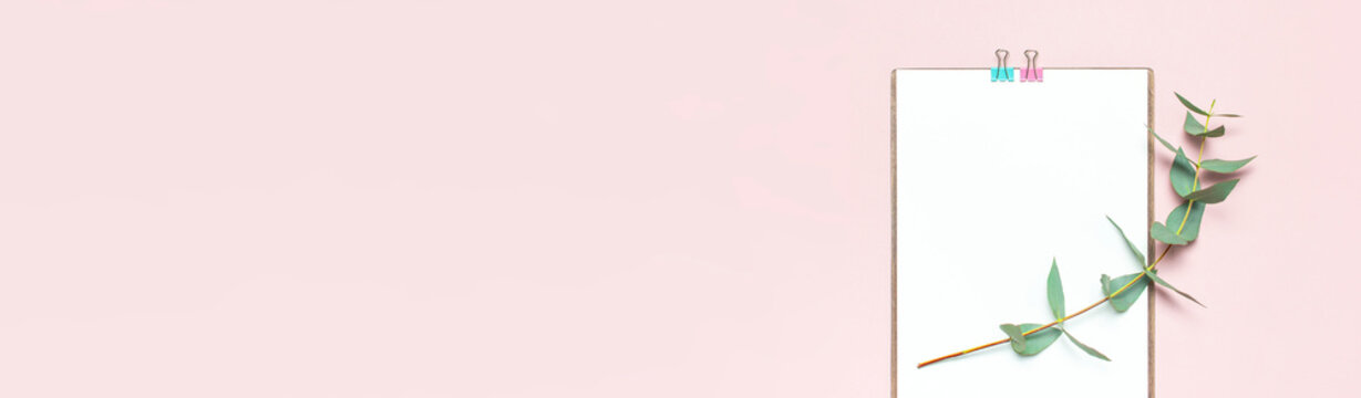 Flat lay clipboard with blank paper, green eucalyptus branches on pastel pink background. Top view female office workspace minimal mock up template concept, copy space. Feminine floral desktop