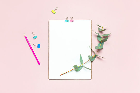 Flat lay clipboard with blank paper, colour pencils, clips, eucalyptus branches on pink background. Top view female office workspace minimal mock up template concept copy space Feminine floral desktop