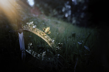 mysterious and magical photo of silver king crown and sword in the England woods or field landscape with light flare. Medieval period concept.