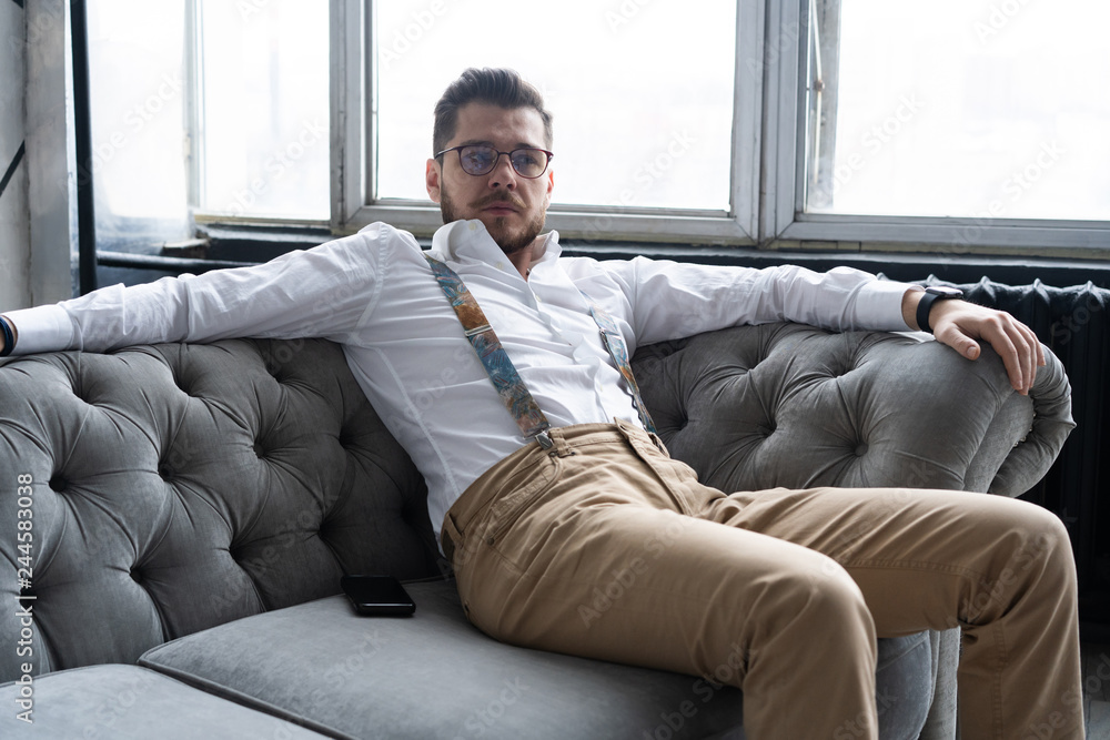 Wall mural enjoying free time. thoughtful young man in full suit looking away while sitting on the sofa at home