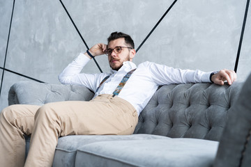 Enjoying free time. Thoughtful young man in full suit looking away while sitting on the sofa at home.