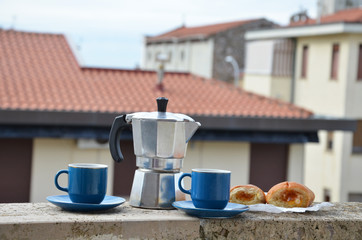 Coffee maker moka pot with roasted coffee beans. Space for text, horizontal. Breakfast concept. Pot and two blue cups of coffee on the balcony with a croissant. On the background of the city.