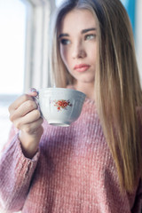 Portrait of a young woman standing next to a window while holding a cup of tea