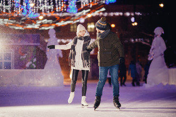 Winter skates, loving couple holding hands and rolling on rink. Illumination in background, night....