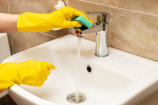 Cleaning washbasin, cleaning service concept. Woman's hands in yellow protective rubber gloves with green sponge under running water from a stainless steel tap on a white ceramic hand basin.