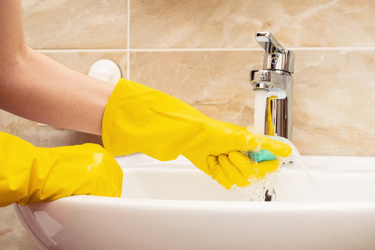 Cleaning washbasin, cleaning service concept. Woman's hands in yellow protective rubber gloves with green sponge under running water from a stainless steel tap on a white ceramic hand basin.