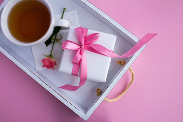 Gift with pink bow and tea in the cup on the white wooden tray on the light pink  background.Top...