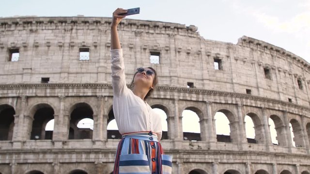 Young woman in fashion dress taking selfies in front of Colosseum in Rome at sunset with smartphone.