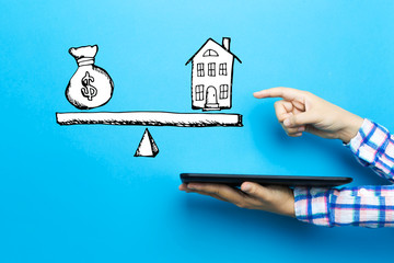 House and money on the scale with a tablet computer on a blue background