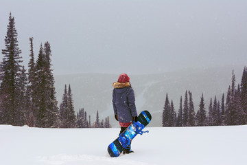 stylish snowboarder girl stand on top of mountain turning back to camera holding her snowboard and...