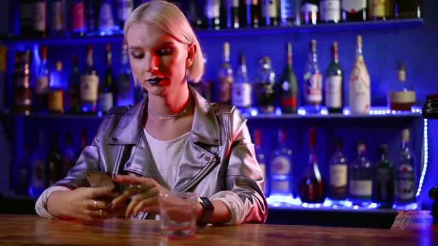 portrait of a young homosexual blonde man in a night bar. drink strong alcohol alone. mobile device for Internet access