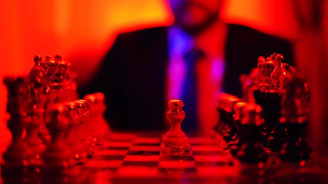 Business man in a business suit sitting on a blurred background plays chess isolated over bright red light background.