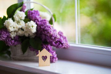 Wooden house with hole in form of heart and tender bouquet of beautiful lilac in white basket near window in daylight.