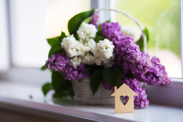 Wooden house with hole in form of heart and tender bouquet of beautiful lilac in white basket near window in daylight.