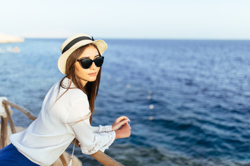 Young beauty woman in sunglasses and hat, cute summer dress standing in feminine carefree pose at the balcony with beautiful sea scenery, enjoying landscape.