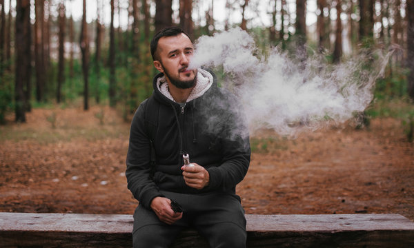 man vaping electronic cigarette in nature. man with beard sit on bench in forest
