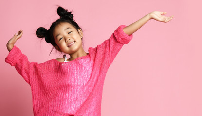 Asian kid girl in pink sweater, white pants and funny buns stands with hands up and smiles. close up - 244602241