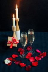 Two glasses with white champagne, gift box and petals of red roses on the background of candles