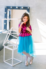 Portrait of little fashion kid girl in a pink shiny blouse and a blue skirt on gray loft background