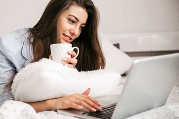 Young smiling woman working on laptop in her bedroom. Happy morning.