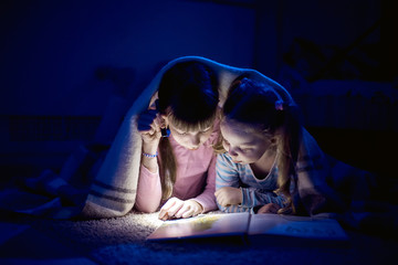 two sisters reading a book with a flashlight in a dark room at night - obrazy, fototapety, plakaty